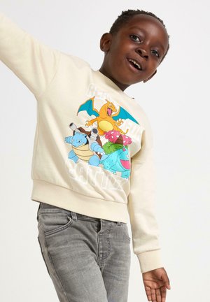 Felpa crema con personaggi cartoon dei Pokémon stampati sul davanti. Il design presenta colori vivaci e polsini a coste. Indossata con jeans grigi.