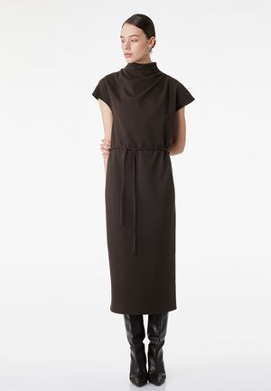 Frau in einem ärmellosen, dunkelbraunen Midikleid mit taillierter Taille und hohem Kragen, trägt schwarze kniehohe Stiefel, steht mit verschränkten Armen hinten.