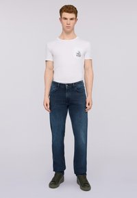 Camiseta blanca de manga corta con un bolsillo en el pecho, combinada con jeans de mezclilla azul oscuro y zapatillas verdes. Fondo liso.