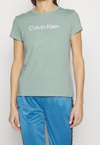 Camiseta de algodón verde claro con mangas cortas y cuello redondo, que presenta el logo de "Calvin Klein" en texto blanco en negrita en el pecho.