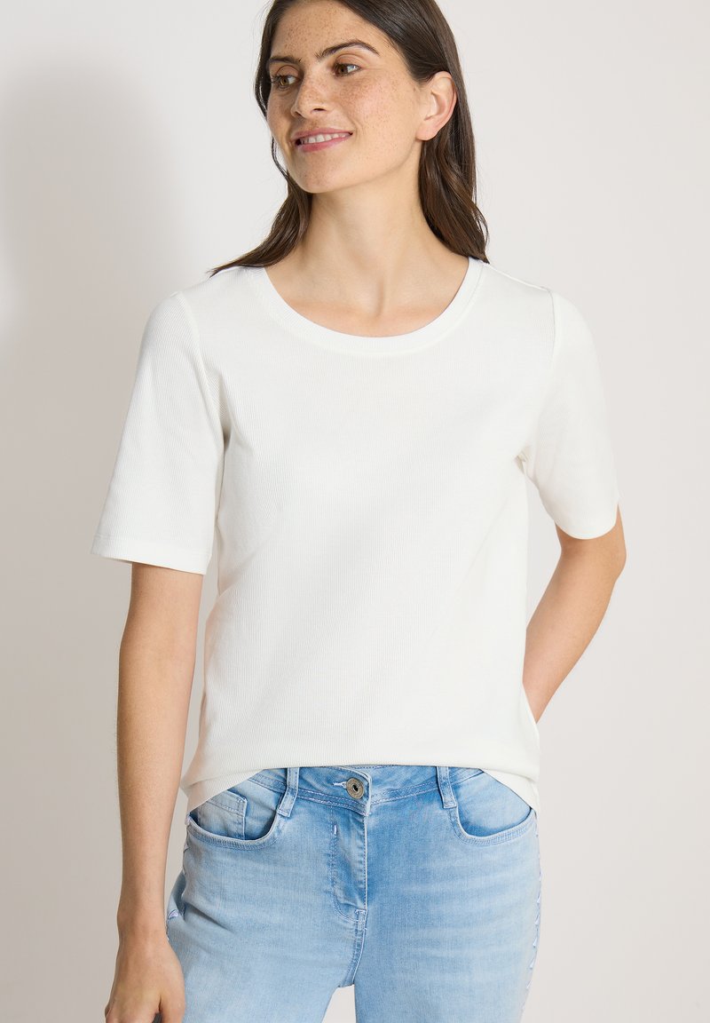 Femme aux longs cheveux foncés portant un t-shirt blanc uni à manches courtes et un jean bleu clair, regardant sur le côté avec un léger sourire.