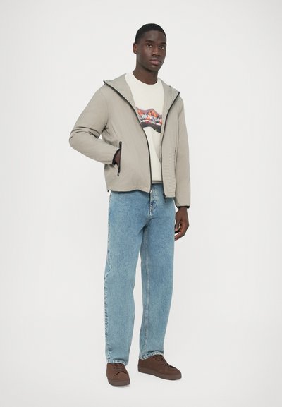 Helehall zip-up jakk tekstuurse kangaga, kantud kreemja trükitud T-särgi kohal, kombineeritud helesiniste teksade ja pruunide vabaaja jalanõudega.