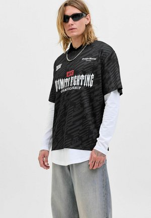 Black graphic T-shirt with a loose fit, featuring a UFC logo and textured pattern; worn over a white long-sleeve shirt and paired with gray jeans.