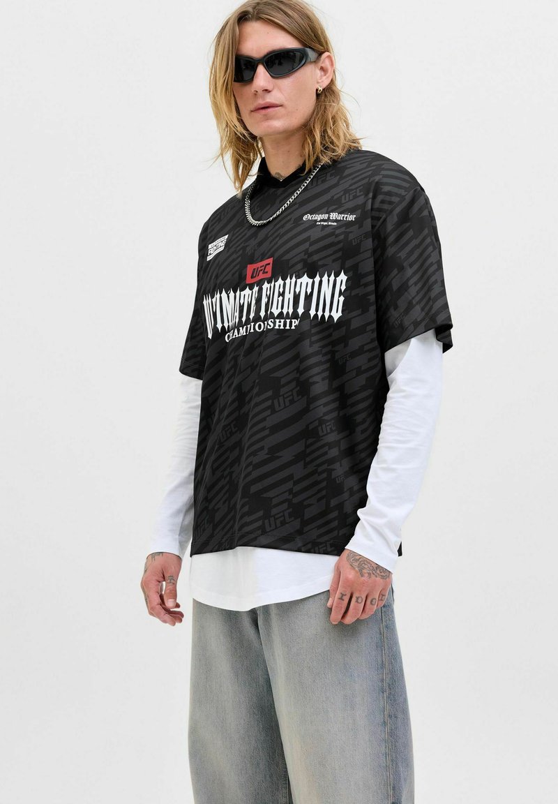 T-shirt graphique noir à coupe décontractée, avec un logo UFC et un motif texturé ; porté par-dessus une chemise à manches longues blanche et associé à un jean gris.