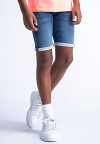 Pantalones cortos de denim azul con puños enrollados, combinados con zapatillas blancas y calcetines blancos. El fondo es blanco liso.