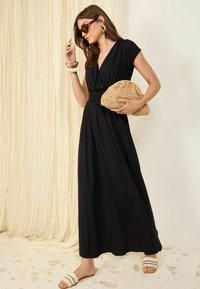 Rochie maxi neagră cu decolteu în V și mâneci scurte, având talie elastică. Modelul ține o poșetă din paie și poartă ochelari de soare și sandale.