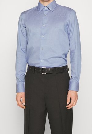 Mężczyzna w jasnoniebieskiej teksturowanej koszuli z długim rękawem schowanej w czarne spodnie z czarnym paskiem, stoi na jednolitym tle.