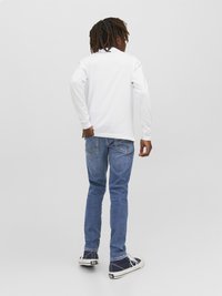 Camisa branca de manga longa combinada com jeans azul e ténis azul-marinho, vista pela parte de trás. Jeans de corte slim com bolsos traseiros, tecido suave.