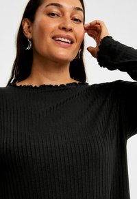 Zwarte geribbelde langemouwen top met een scalloped halslijn en textuurstof. Gepresenteerd met een model tegen een neutrale achtergrond.