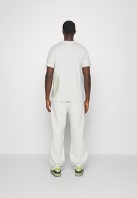 Lys grå kortærmet T-shirt og hvide sweatpants båret af en person, med et rundt logo detajle på skjortens bagside og sko med grønne accenter.