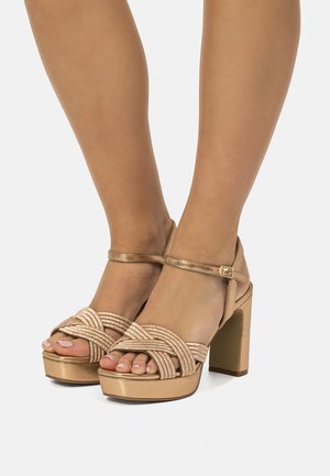 Piernas que llevan sandalias de plataforma de tacón alto en metalizado dorado con tiras frontales trenzadas y cierres de hebilla en el tobillo, sobre un fondo blanco.