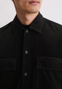 Uomo che indossa una camicia in velluto a coste nera con grandi tasche anteriori e colletto abbottonato, mostrato dalla gola al petto.