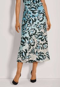 Florale Midirock mit blauen, schwarzen und weißen Blumenmustern auf hellem Hintergrund. Glattes Material, ausgestellte Form, getragen mit schwarzen High Heels.