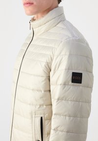Gros plan d’une personne portant une veste matelassée beige clair avec une fermeture éclair noire et un patch logo BOSS sur la manche supérieure.