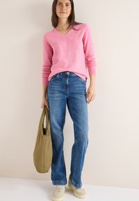Hellrosa V-Ausschnitt-Pullover, blaue gerade Jeans, beiger Beutel und cremfarbene Sneaker. Weicher Stoff mit gerippten Bündchen und Saum.