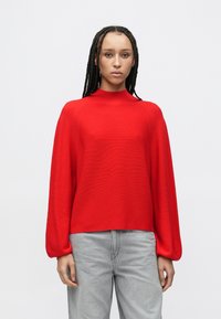 Czerwony sweter z dzianiny z wysokim kołnierzem, szerokimi rękawami i teksturowanym wykończeniem. Połączony z jasnoszarymi dżinsami.