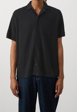Person iført en løs, sort knap-op kortærmet skjorte med brystlomme og mørkeblå jeans mod en ensfarvet baggrund.