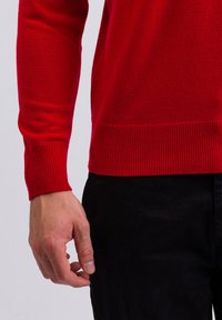 Maglione rosso a maglia con polsini e orlo a coste, che mostra una texture liscia. La mano del modello è appoggiata casualmente vicino alla vita, indossando pantaloni neri.