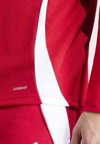 Red athletic shirt with white accents, featuring an "AEROREADY" logo. Smooth fabric texture, close-fitting design, highlighting stitching details.