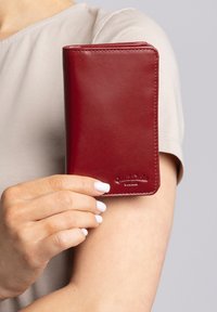 Červená kožená peňaženka s hladkou textúrou, zaoblenými rohmi a vyrazenou značkou. Držená v ruke na neutrálnej pozadí.