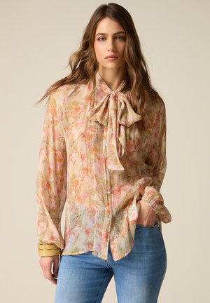 Donna che indossa una blusa trasparente a fiori con colletto a fiocco e jeans blu, in piedi con una mano in tasca davanti a uno sfondo uniforme.