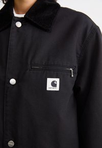 Veste Carhartt noire avec col en velours côtelé, boutons-pression argentés, poche zippée sur la poitrine et écusson logo sur fond blanc.