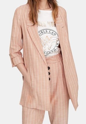 Vrouw draagt een lichtroze fijn gestreepte blazer en bijpassende hoge taille broek met een wit grafisch T-shirt in de broek gestopt.