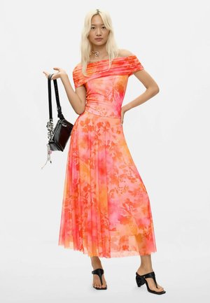 Vrouw met lang blond haar die een oranje-roze off-shoulder bloemenjurk draagt, een zwarte handtas vasthoudt en zwarte sandalen met hakken draagt.