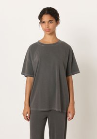 Mlada ženska z kodrastimi lasmi, ki nosi ohlapno temno sivo majico in ujemajoče hlače, stoji pred enostavnim belim ozadjem.