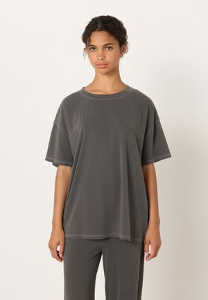 Junge Frau mit lockigem Haar, die ein locker sitzendes dunkelgraues T-Shirt und passende Hose trägt, vor einem schlichten weißen Hintergrund stehend.