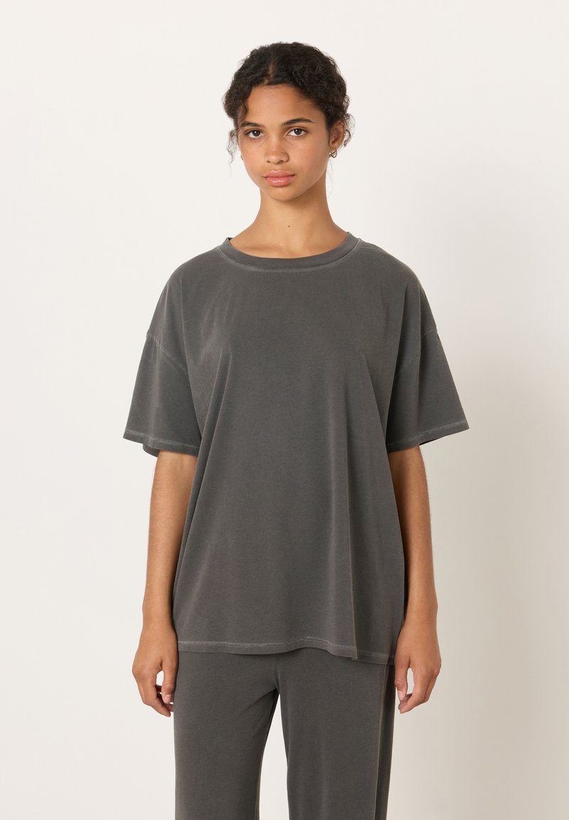 Mlada ženska z kodrastimi lasmi, ki nosi ohlapno temno sivo majico in ujemajoče hlače, stoji pred enostavnim belim ozadjem.