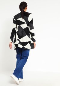 Cardigan lungo a righe bianche e nere con una maglia morbida e testurizzata, caratterizzato da forme geometriche, apertura frontale e maniche lunghe. Abbinato a jeans blu.