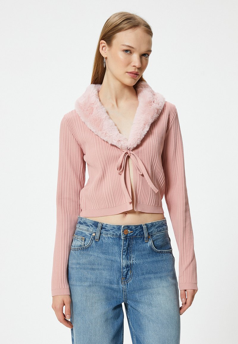 Rosa gerippter kurzer Cardigan mit einem Kunstfellkragen, mit einer Bindeschließe vorne und langen Ärmeln, kombiniert mit hochgeschnittenen blauen Jeans.