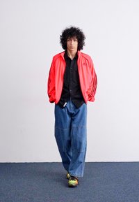 Red nylon tracksuit jacket with black shirt, oversized blue denim pants, and colorful sneakers, featuring contrasting stripes and relaxed fit.
