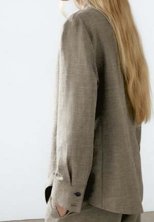 Personne aux longs cheveux blonds portant un blazer marron clair texturé avec les manches boutonnées aux poignets, debout devant un fond blanc uni.