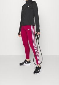 Schwarzes Sportoberteil mit langen Ärmeln, bordeauxrote Leggings mit weißen Streifen und schwarze Sneakers. Hält ein schwarzes Springseil in der Hand.