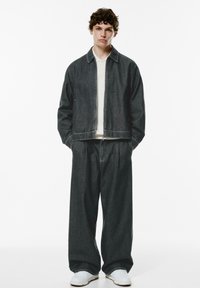 Mørk denimjakke og brede bukser med en afslappet pasform. Lysfarvet skjorte under og hvide sneakers fuldender outfittet.