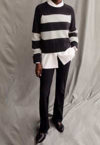 Musta-valkoraidallinen neulepaita pyöreällä kaula-aukolla, kerrospukeutunut valkoisen paidan päälle. Yhdistetty mustiin housuihin ja kapeakärkisiin mustiin kenkiin.