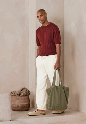 Vyras dėvi bordo spalvos trumpomis rankovėmis marškinį, baltas kelnes, smėlio spalvos slip-on batus, laiko didelę alyvuogių žalios spalvos rankinę, stovi viduje šalia krepšelio ir pagalvėlės.