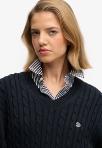 Navy blue cable-knit sweater with a V-neck, layered over a white and navy striped collared shirt, featuring a small embroidered logo.