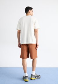 Conjunto casual que incluye una camiseta de algodón color crema, pantalones cortos marrones y zapatillas grises con detalles amarillos. Ajuste simple y relajado, sin patrones.