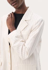 Blazer blanco a rayas con un diseño ajustado, que presenta solapa muesca, un bolsillo en el pecho y puños abotonados. Textura de tela suave.