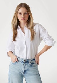 Camicia bianca con bottoni, dal taglio morbido e maniche arrotolate, con una trama sottile. Abbinata a jeans in denim azzurro chiaro, leggermente strappati.