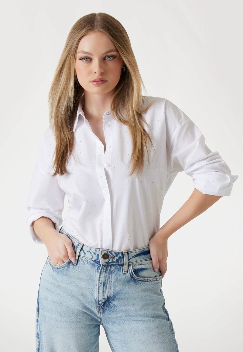 Camicia bianca con bottoni, dal taglio morbido e maniche arrotolate, con una trama sottile. Abbinata a jeans in denim azzurro chiaro, leggermente strappati.