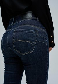 Jeans de mezclilla azul oscuro con un corte ajustado, que presentan sutiles detalles de costura y una etiqueta de cuero negro con la marca en la parte posterior de la cinturilla.
