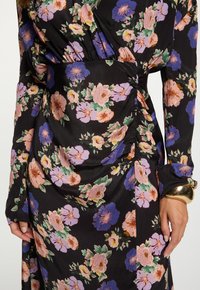 Zwart jurkje met een roze en paarse bloemenprint, lange mouwen en eenGathered taille detail, gedragen met een grote gouden armband om de pols.