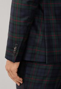 Primo piano di una mano accanto a una manica di un completo tartan a quadri verde scuro e blu navy, con quattro bottoni e pantaloni abbinati.