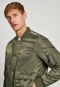Chaqueta bomber verde oliva con una textura brillante y acolchada. Cuenta con bolsillos delanteros, un bolsillo con cremallera en la manga y puños acanalados.