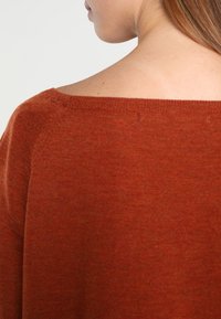 Femme portant un pull couleur rouille, à col bateau, montrant le dos et l'épaule droite, avec une peau claire et des cheveux bruns lisses.