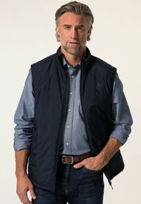 Marinblå väst av lättviktig tyg, med dragkedja fram, sidofickor, bärs över en ljusblå skjorta med knappar, tillsammans med mörk denimjeans.